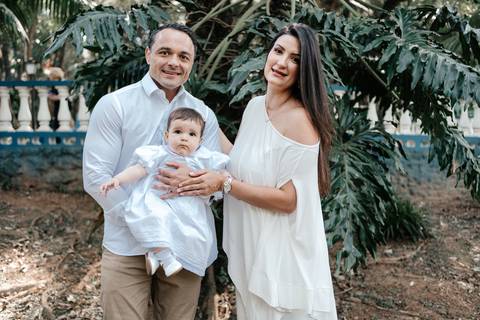 Foto do casal sorridente em pé na área externa segurando a bebê no colo.'