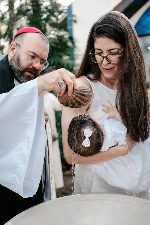 Foto do momento exato do batismo, com o padre derramando a água na cabeça da bebê com uma concha.'