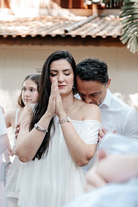 Retrato comovente da mãe de olhos fechados com as mãos em prece, recebendo o carinho do pai.'