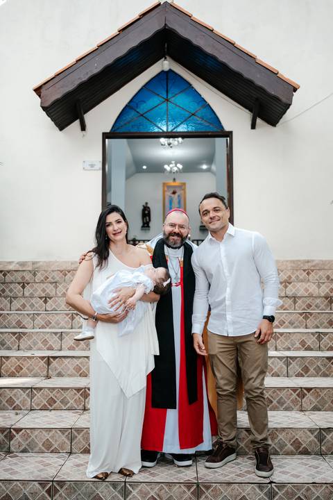 Retrato do casal com a bebê no colo posando ao lado do padre nos degraus da igreja.'