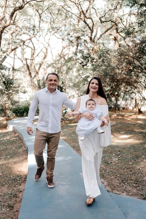 Foto do casal caminhando de mãos dadas pelo jardim da igreja com a bebê.'
