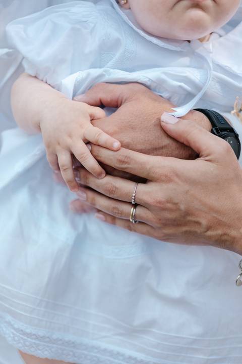 Foto em detalhe das mãos dos pais envolvendo a mãozinha delicada da bebê.'