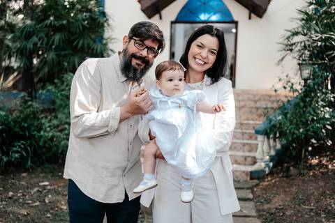 Retrato da bebê no colo dos padrinhos sorridentes na área externa da igreja.'