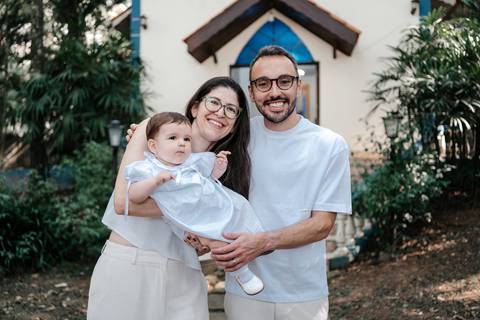 Retrato da bebê no colo de casal de padrinhos vestindo tons claros na escadaria.'