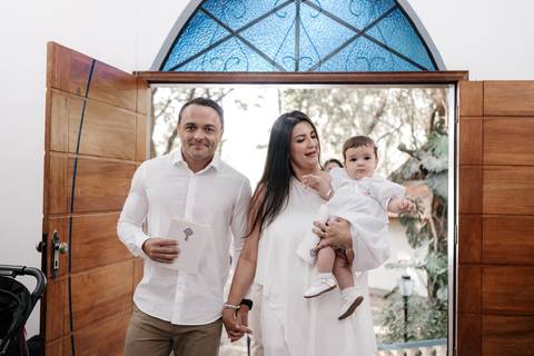 Foto do casal entrando na capela; o pai segura a vela batismal e a mãe carrega a bebê.'