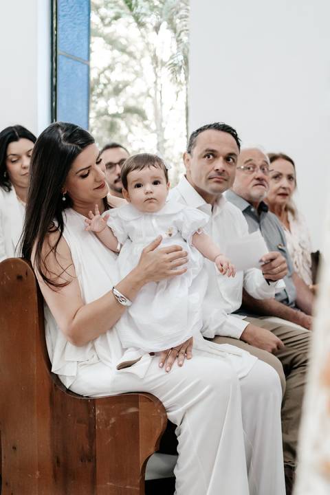 Foto aproximada da mãe segurando a bebê no colo atentamente junto aos familiares no banco.'