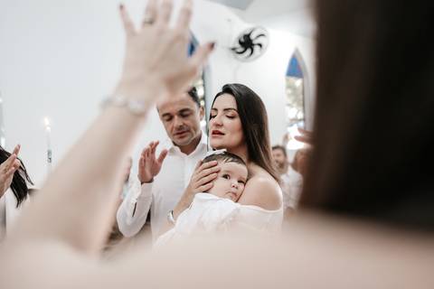 Foto em ângulo contra-plongê focando nos pais com a bebê no colo durante as preces.'