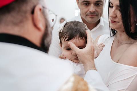 Foto do padre ungindo a testa da bebê com o óleo sagrado sob o olhar atento dos pais.'