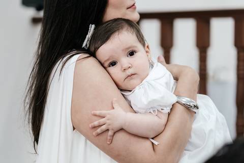 Foto aproximada da mãe abraçando firmemente a bebê no colo em momento de devoção.'