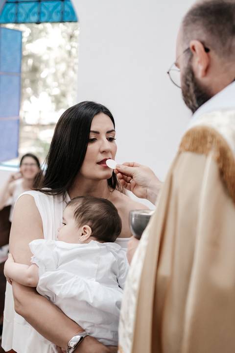 Foto da mãe recebendo a comunhão (eucaristia) do padre enquanto segura a bebê.'