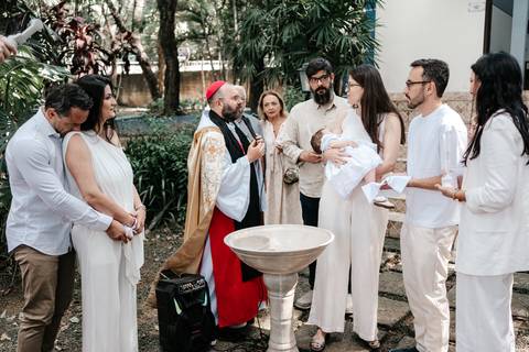 Foto panorâmica da família e do clero reunidos ao redor da pia batismal na área externa.'