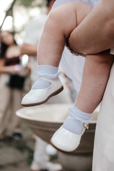 Detalhe em close do sapatinho de verniz branco e da meia com renda da bebê na pia batismal.'