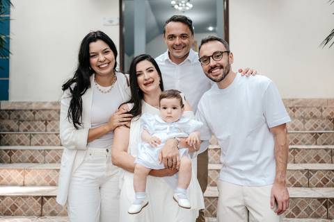 Retrato da bebê com os padrinhos de batismo sorridentes, todos vestindo branco.'