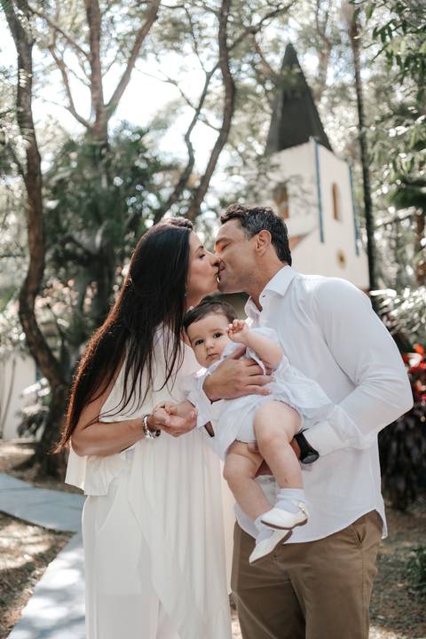 Retrato romântico do casal trocando um beijo enquanto a mãe segura a bebê.'