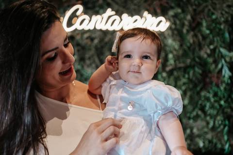 Foto da mãe com a bebê na recepção, com painel decorado e letreiro iluminado ao fundo.'