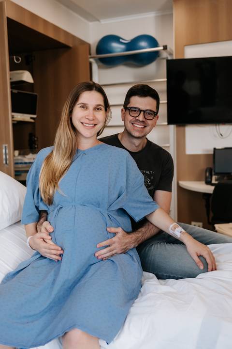 Retrato do casal sorridente no quarto da maternidade com o pai abraçando a gestante por trás.'