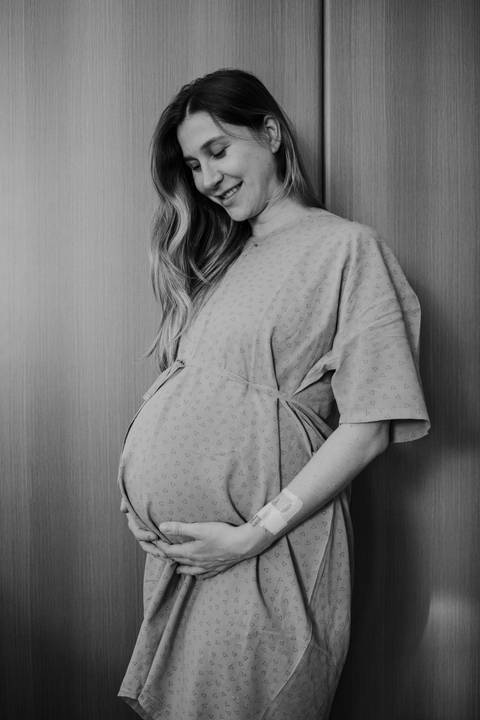 Foto em preto e branco da gestante segurando a barriga no quarto do hospital.'