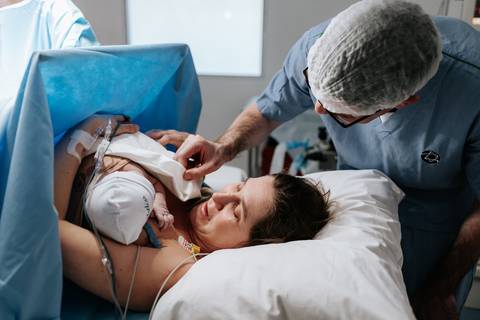 Foto da mãe segurando o bebê envolto em campos cirúrgicos, acompanhada pelo parceiro e equipe.'