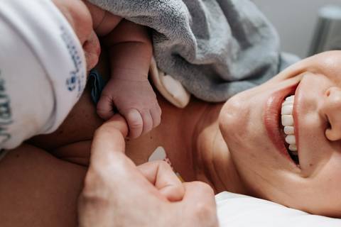 Close do sorriso radiante da mãe com foco no toque delicado da mãozinha do bebê em seu peito.'