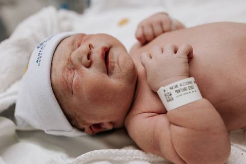 Foto do bebê Mathias deitado de costas, usando touquinha e pulseira de identificação hospitalar.'