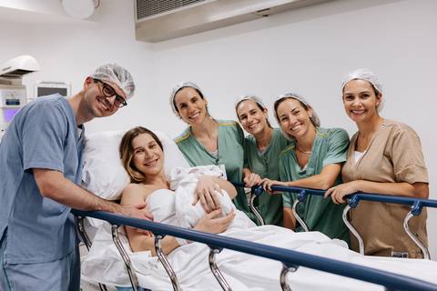 Retrato oficial do casal com o bebê no colo, cercados pela equipe médica na sala cirúrgica.'