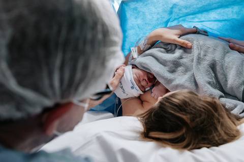 Foto do parceiro observando emocionado a mãe e o recém-nascido aninhados na mesa de parto.'