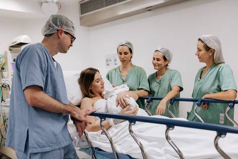 Retrato do casal com o bebê no leito hospitalar, cercados por enfermeiras sorridentes.'