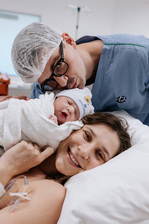 Retrato de close do casal sorridente com o bebê envolto em uma manta branca.'