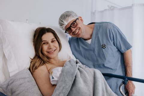 Retrato oficial do casal posando feliz com o filho Mathias no quarto da maternidade.'