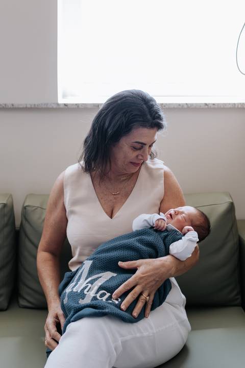 Foto da avó sentada no sofá segurando o neto Mathias no colo com um olhar carinhoso.'