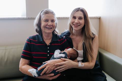 Retrato da avó ao lado da mãe corujando o bebê no quarto do hospital.'