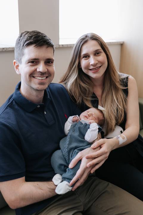Retrato de close do casal sorridente com o bebê Mathias adormecido entre eles.'