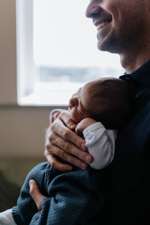 Foto aproximada do pai ninando o bebê contra o peito, com foco no perfil do recém-nascido.'