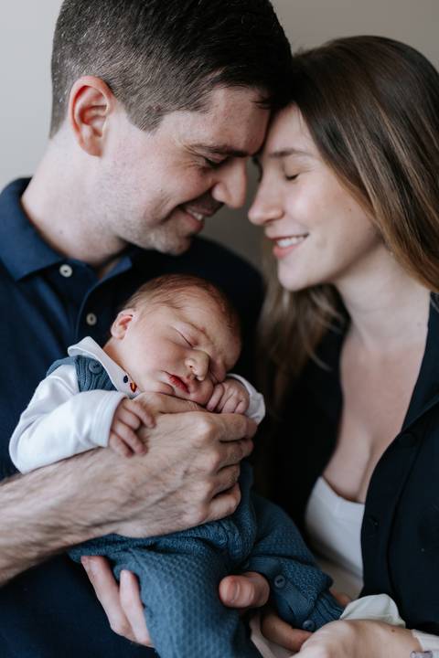 Close do casal sorrindo com os rostos bem próximos ao pequeno Mathias, que dorme no colo.'