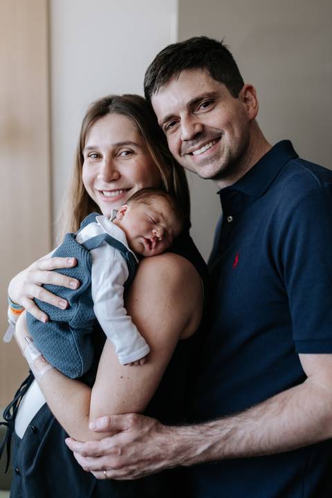Retrato do casal sorridente posando com o bebê Mathias acolhido nos braços da mãe.'