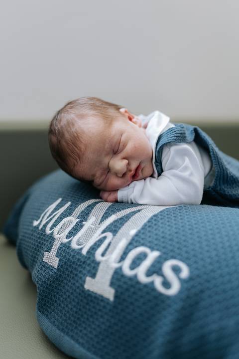 Foto do bebê dormindo de bruços em cima da almofada azul bordada com o nome 