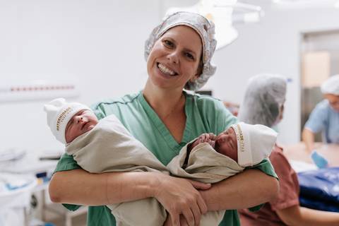 Foto de integrante da médica sorridente segurando os dois gêmeos no colo.'