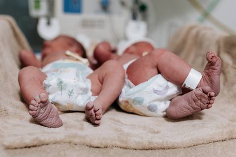 Foto de ângulo aberto dos irmãos gêmeos deitados na manta hospitalar, destacando seus pezinhos.'