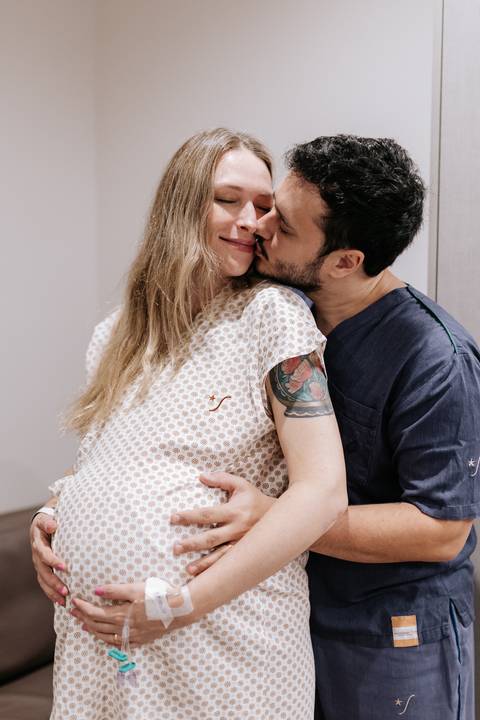 Foto aproximada da gestante recebendo um beijo carinhoso do parceiro no quarto do hospital.'