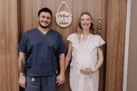 Retrato do casal sorridente em pé ao lado da porta do quarto de maternidade.'