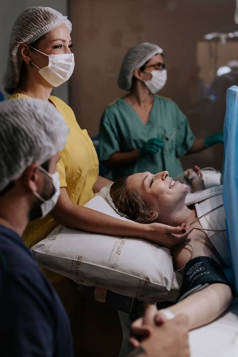 Foto da gestante sorrindo na maca cirúrgica enquanto recebe o carinho do parceiro.'