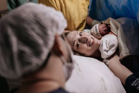 Retrato do casal emocionado na mesa cirúrgica com o primeiro bebê aconchegado no colo.'