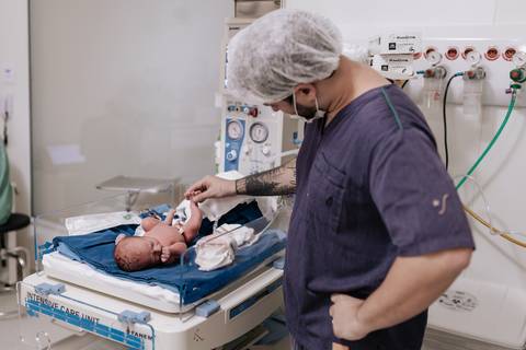 Foto do pai observando o bebê no berço aquecido enquanto a equipe realiza os procedimentos.'