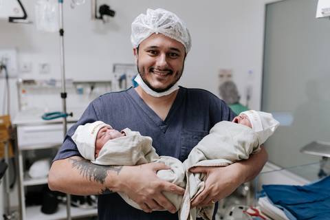 Foto do pai sorridente e orgulhoso segurando os dois bebês gêmeos no colo na sala de parto.'