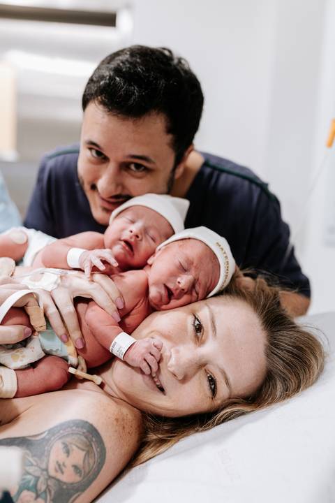 Retrato oficial da família com o casal sorrindo e os dois bebês juntos sobre o peito da mãe.'