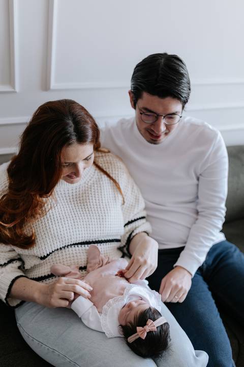 Foto do casal sentado no sofá admirando e sorrindo para a bebê recém-nascida em seu colo.'
