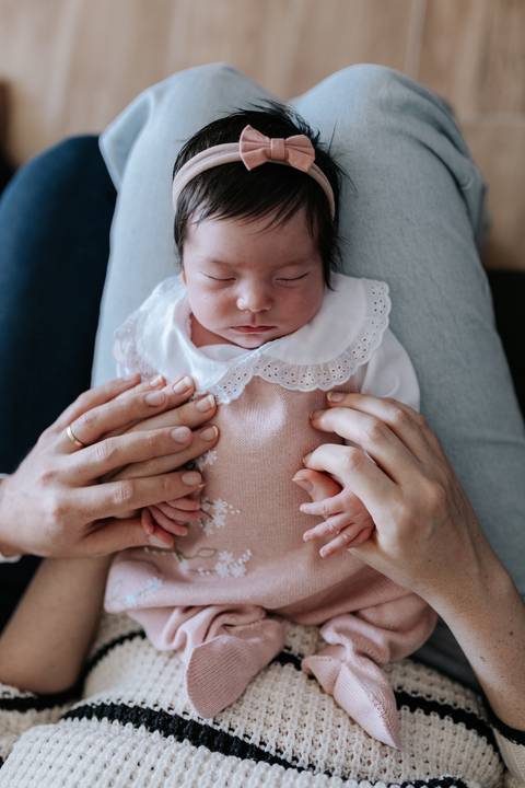 Detalhe das mãos da mãe e do pai amparando delicadamente o corpo da bebê, que veste roupa rosa.'