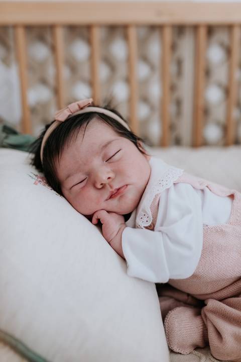 Close da bebê dormindo aconchegada em um travesseiro branco, com a mãozinha perto do rosto.'