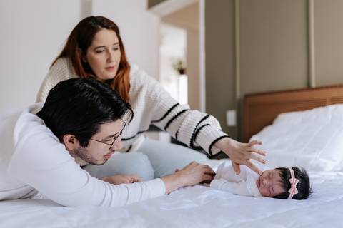 Foto do casal deitado na cama interagindo e acariciando a bebê que está entre eles.'