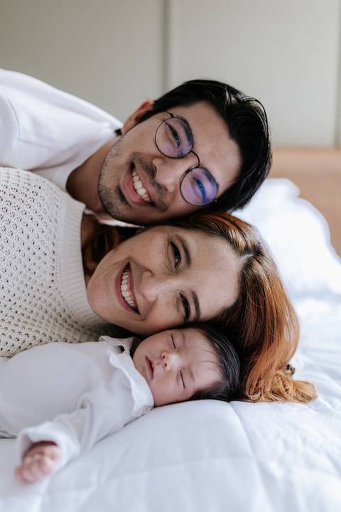 Retrato de close do casal sorridente deitado de bruços bem pertinho da bebê adormecida.'